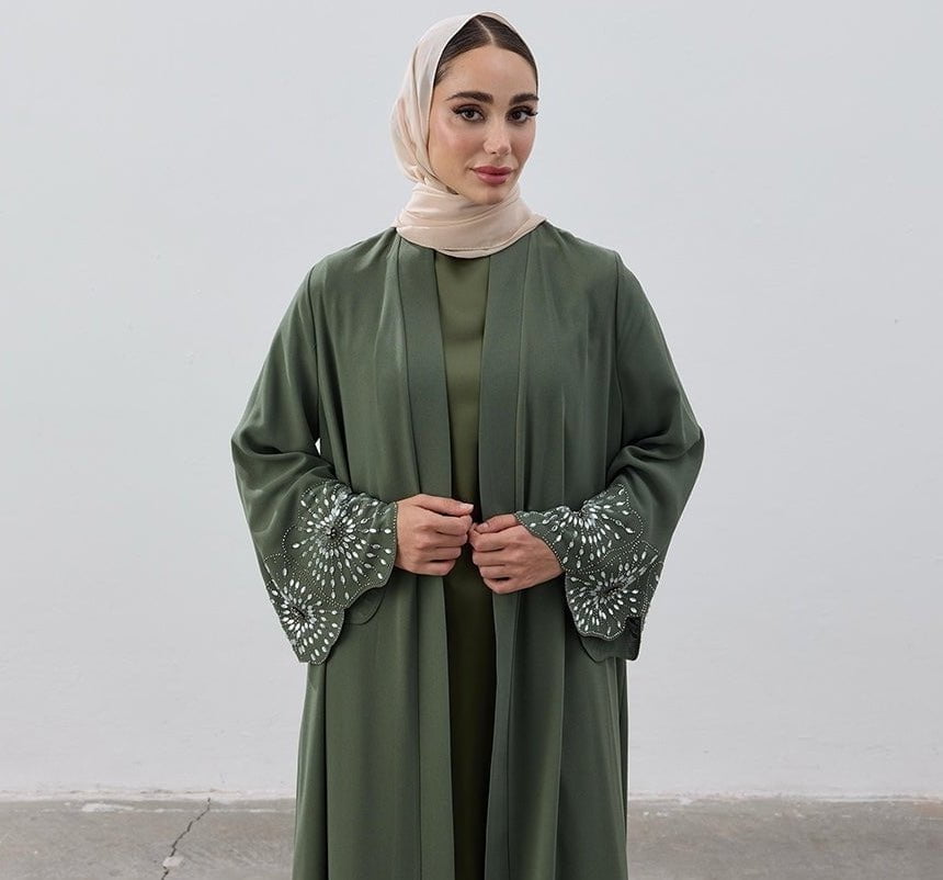 Woman wearing a green abaya with intricate patterns on the sleeves against a plain background